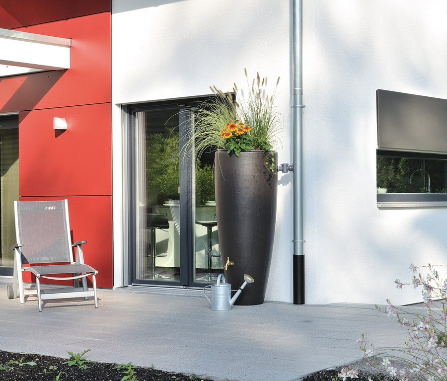 Auf einer Terrasse steht ein braunes 300 l Garantia 2-in-1-Regentonnen-Komplett-Set mit Blumen und einer Gießkanne.