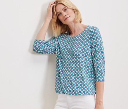 Frau posiert an einer Wand in einem Blusenshirt mit Alloverprint und einer weißen Hose.