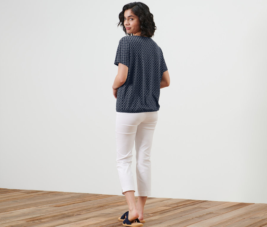 Frau blickt über die Schulter und trägt ein bedrucktes Blusenshirt, weiße Hose und Sandalen.