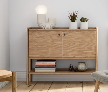 Einrichtung mit einer Anrichte aus Holz, einer LED-Design-Tischleuchte, einem Sessel mit Fußhocker, Topfpflanzen und Dekorationen.