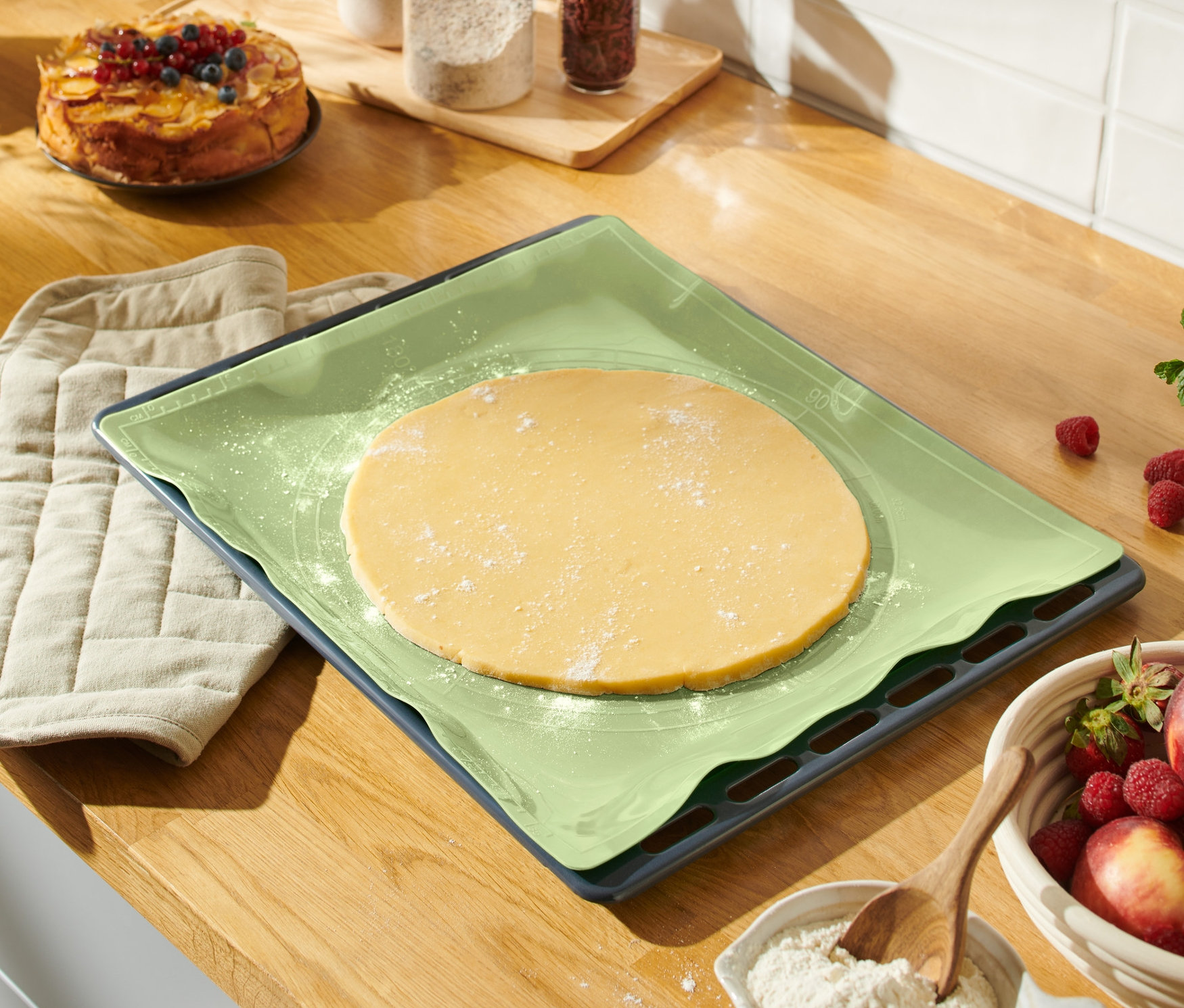 Auf einer Arbeitsfläche liegen eine Dauerbackmatte mit ausgerolltem Teig, ein Topflappen und eine Schüssel mit Obst.