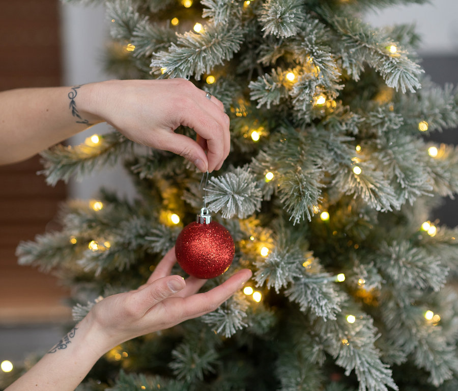 Eine Person schmückt einen künstlichen, beschneiten Weihnachtsbaum "Fichte" mit LED-Lichterkette mit einer roten Kugel.