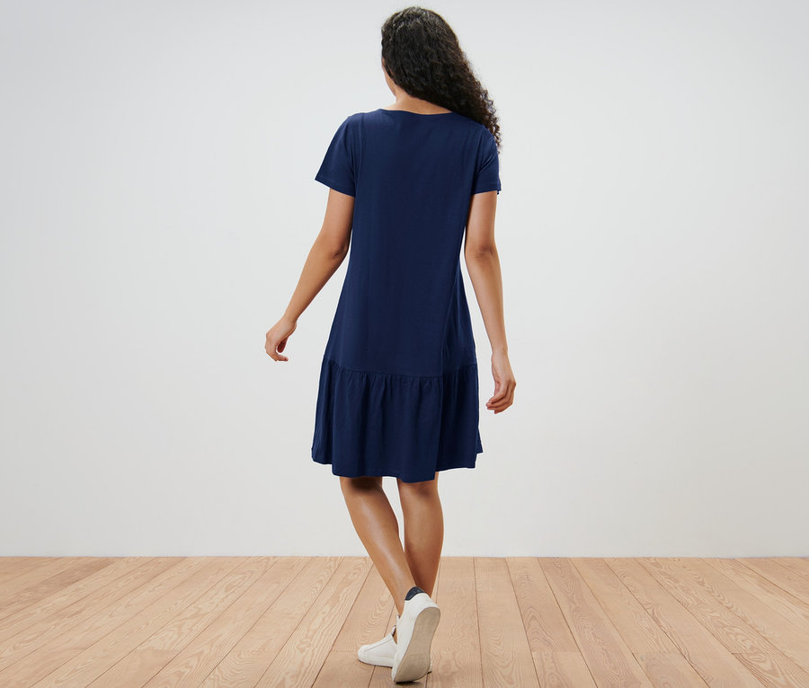 Rückansicht einer Frau in marineblauem Jerseykleid mit Volant und weißen Turnschuhen.