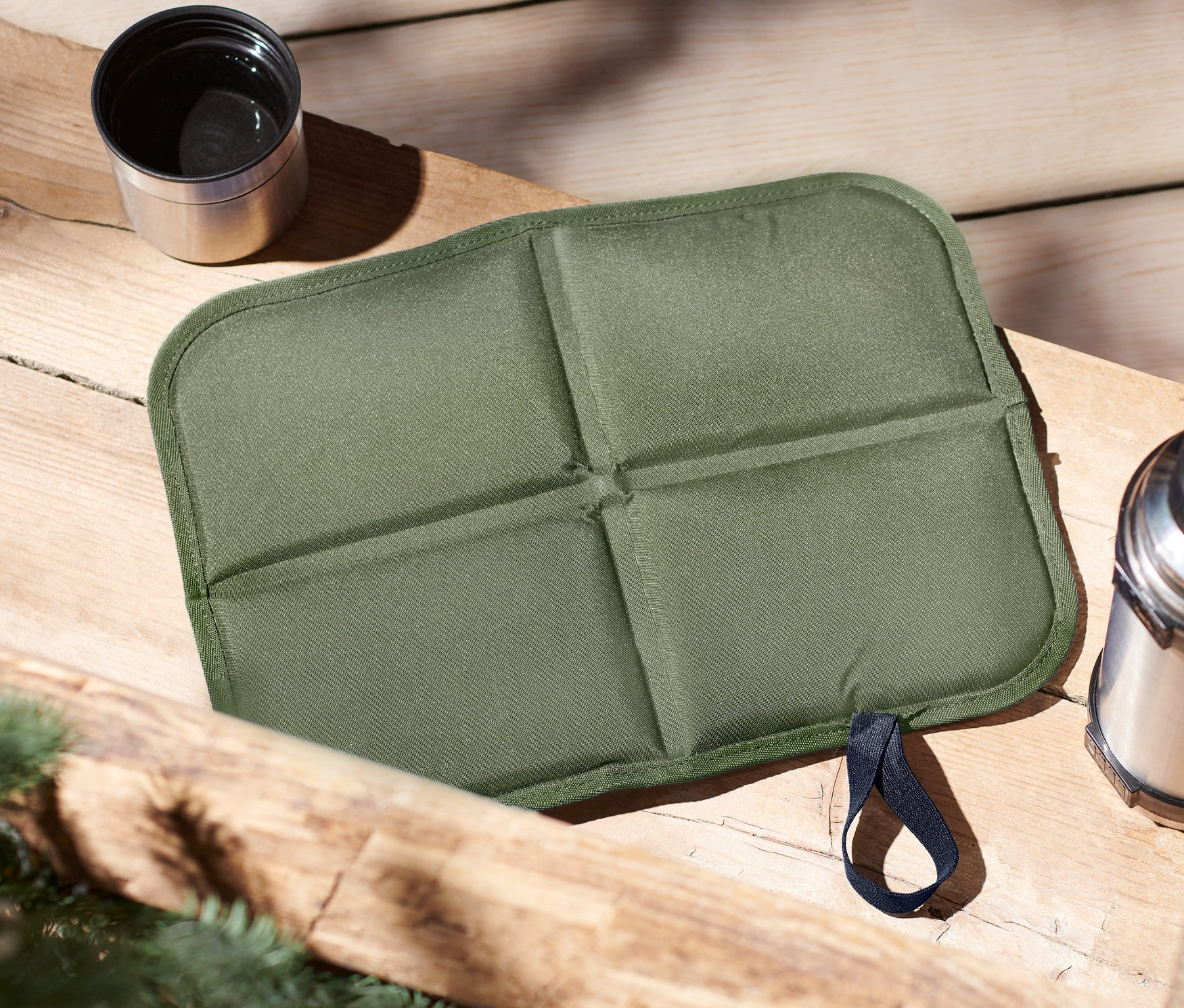 Auf einem Holztisch liegen ein grünes, faltbares Thermo-Sitzkissen, eine Thermoskanne und eine Tasse.