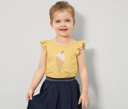 Ein lächelndes kleines Mädchen trägt ein gelbes Kinder-Shirt mit Volantärmeln und einem Eisaufdruck.