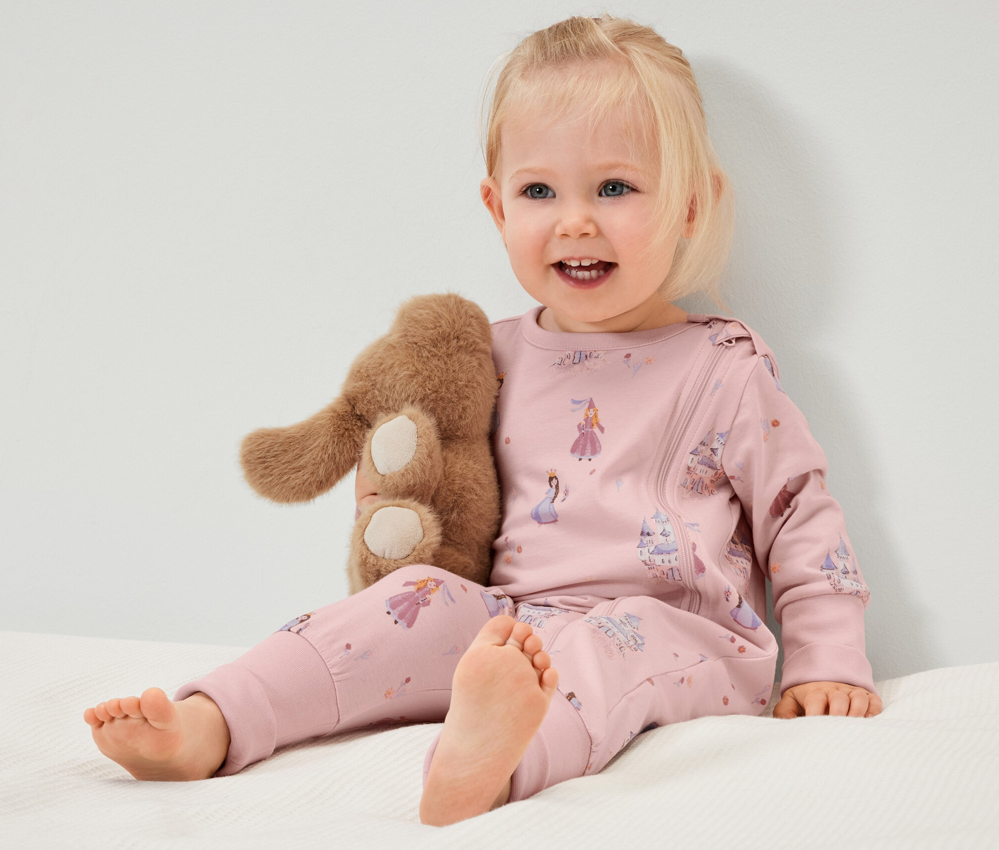 Ein lächelndes Kleinkind sitzt auf einem Bett und umarmt ein Stoffkaninchen. Es trägt einen rosa Baby-Pyjama mit Prinzessinnenmuster.