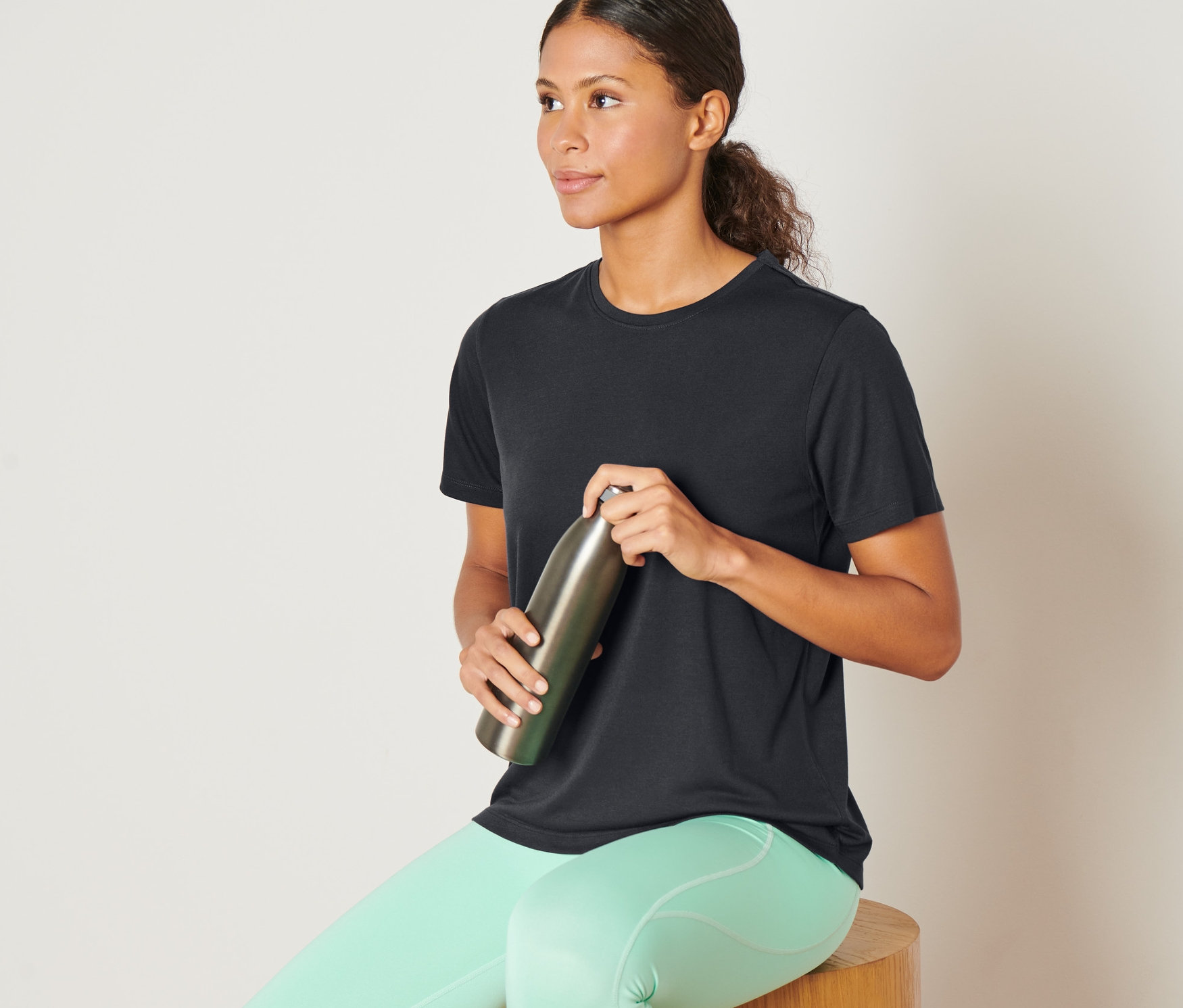 Frau sitzt auf einem Holzhocker und hält eine Metallflasche. Sie trägt ein anthrazitfarbenes Sportshirt.