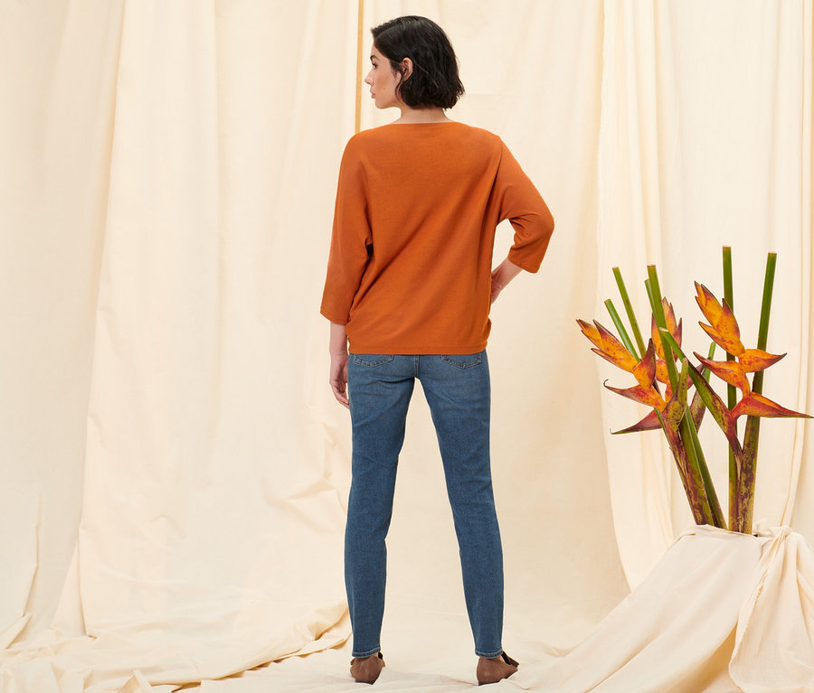 Frau steht mit dem Rücken zur Kamera und trägt eine Slim-Denim »Fit Emma« in Midblue Denim, einen orangefarbenen Feinstrickpullover und Loafer aus Veloursleder.
