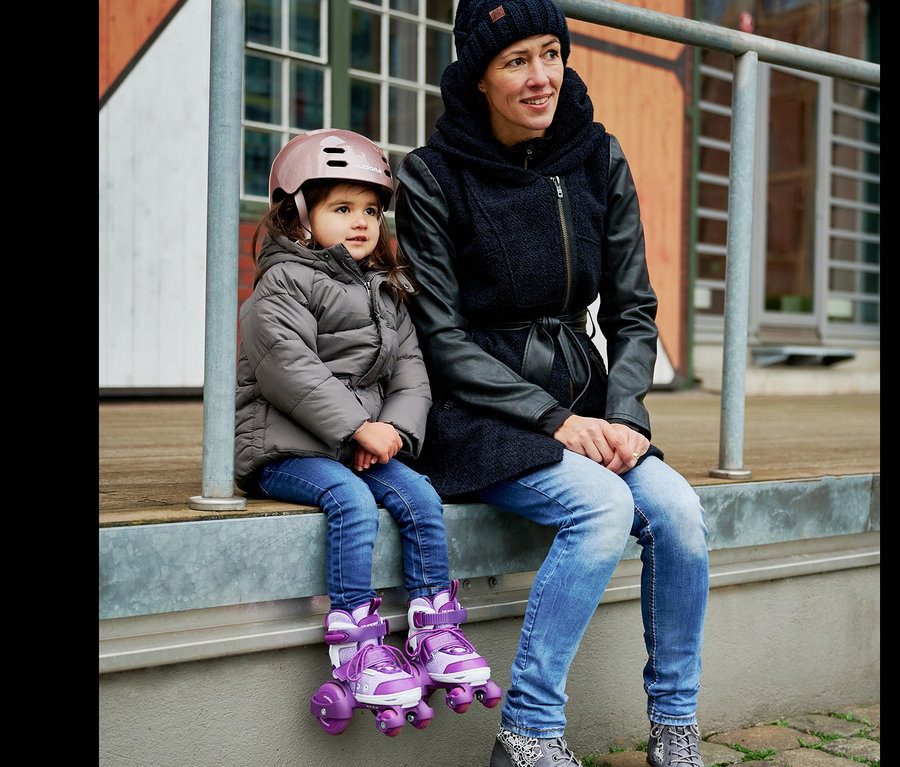 Eine Frau und ein Mädchen sitzen nebeneinander. Das Mädchen trägt einen rosafarbenen Helm und lila HUDORA My First Quad Rollschuhe.