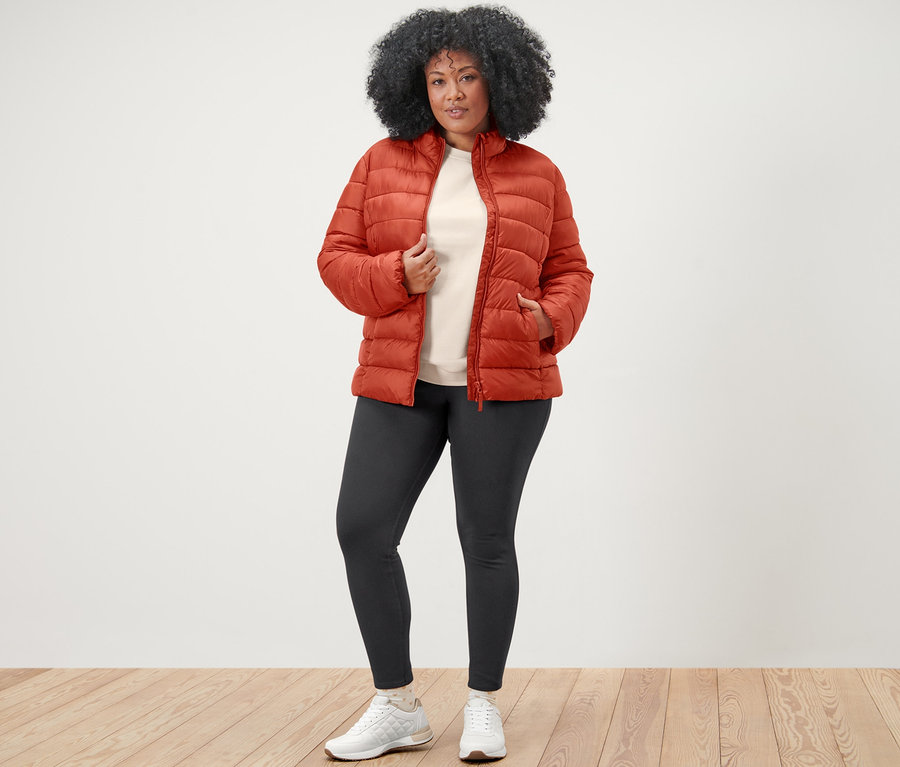 Frau posiert in orangefarbener Steppjacke, Sweatshirt mit Stickerei und Jeggings.