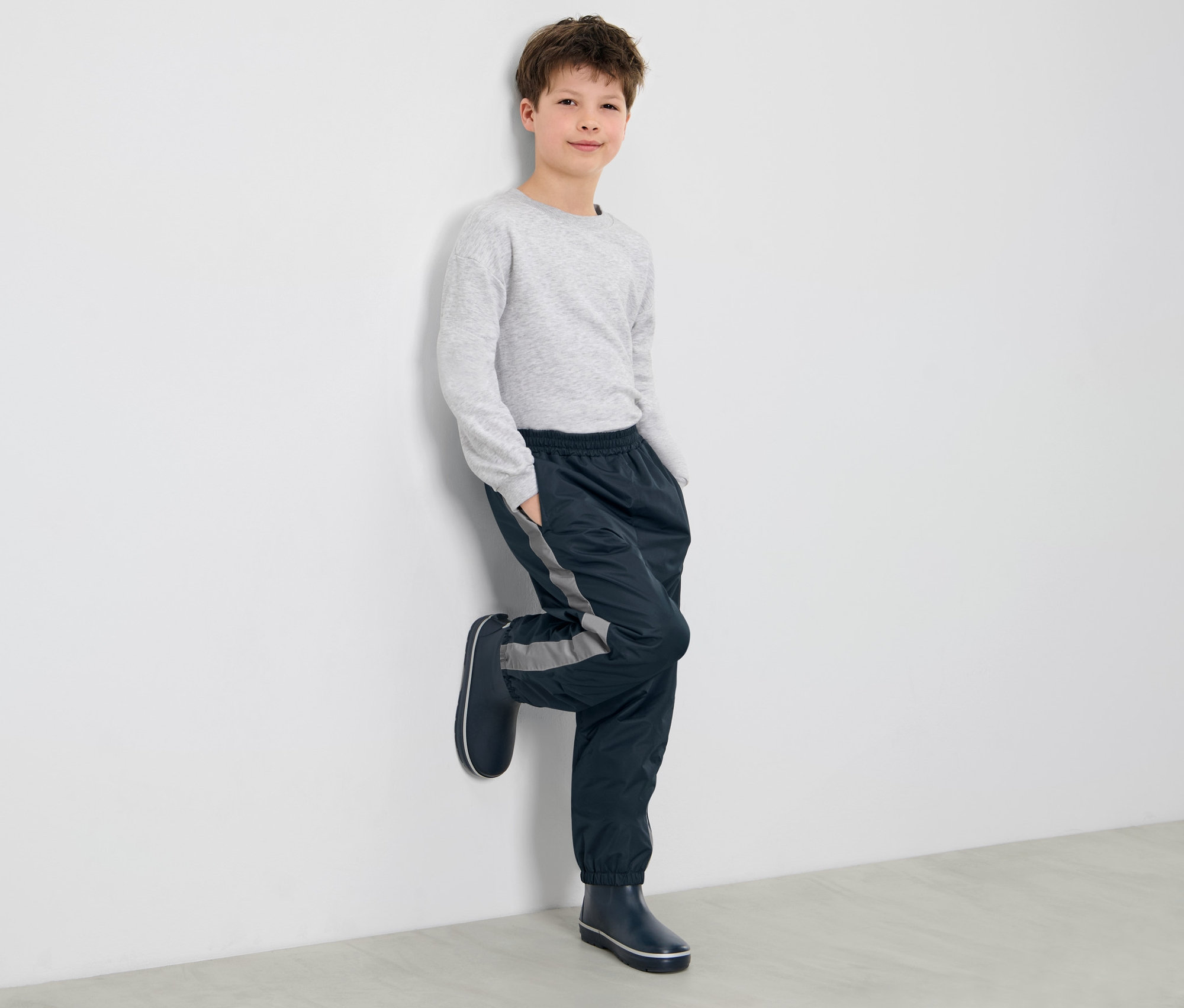 Ein Junge lehnt an einer Wand und trägt eine Kinder-Regenhose und Gummistiefel.