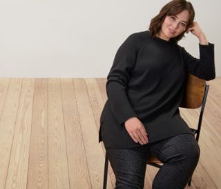 Frau sitzt auf einem Stuhl und trägt einen langen schwarzen Strickpullover und eine bedruckte Bengalin-Hose.