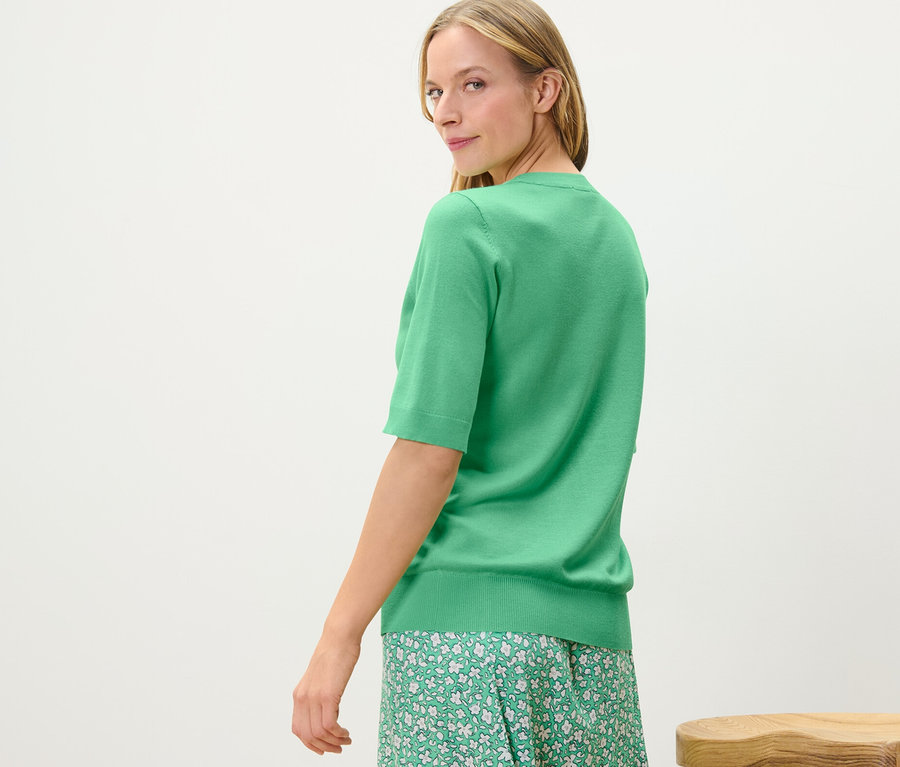 Eine Frau blickt über die Schulter und trägt einen grünen Feinstrickpullover und einen geblümten Rock.