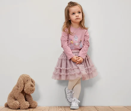 Ein Mädchen in einem rosa Langarmshirt mit Prinzessinnenmotiv, einem rosa Rock und weißen Turnschuhen steht an einer Wand. Neben ihr sitzt ein Plüschhund.