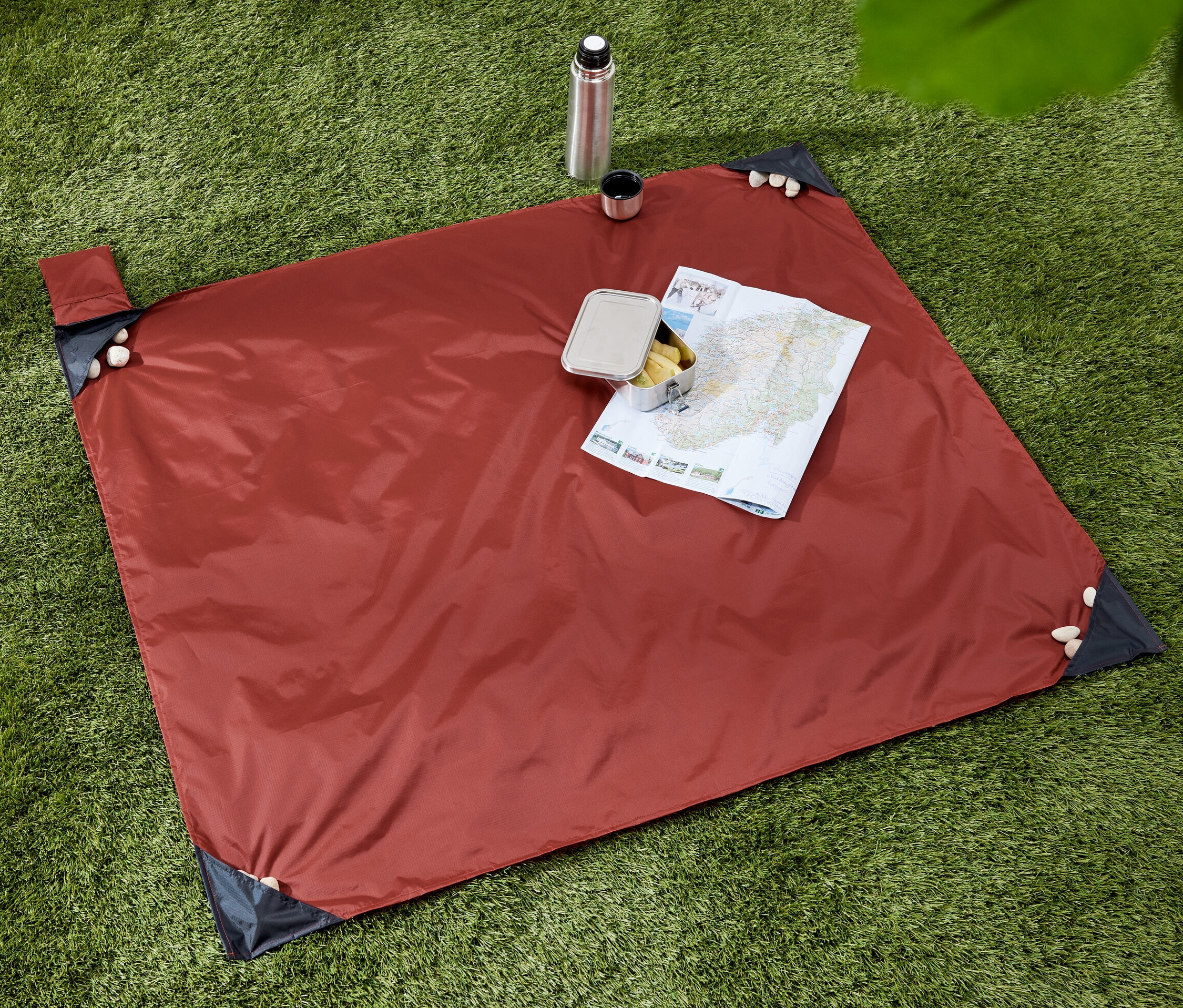 Auf grünem Gras liegt eine Mini-Picknickdecke. Auf der Decke liegen eine Landkarte und eine Brotbox. Eine Thermoskanne mit Becher steht daneben.