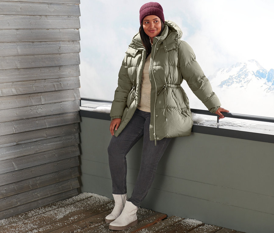 Eine Frau lehnt an einem Geländer und trägt eine Daunenjacke, Slimfit-Jeans, Veloursleder-Boots und eine bordeauxrote Strickmütze.