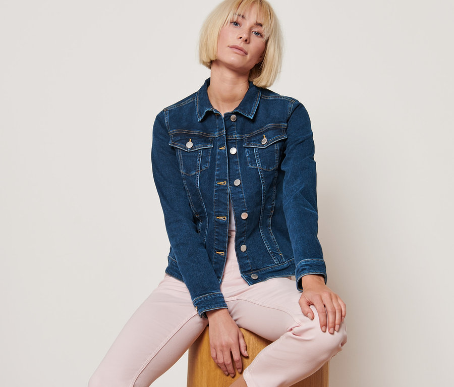 Frau sitzt auf einem Holzhocker und trägt eine Jeansjacke in Mid-Blue-Denim.