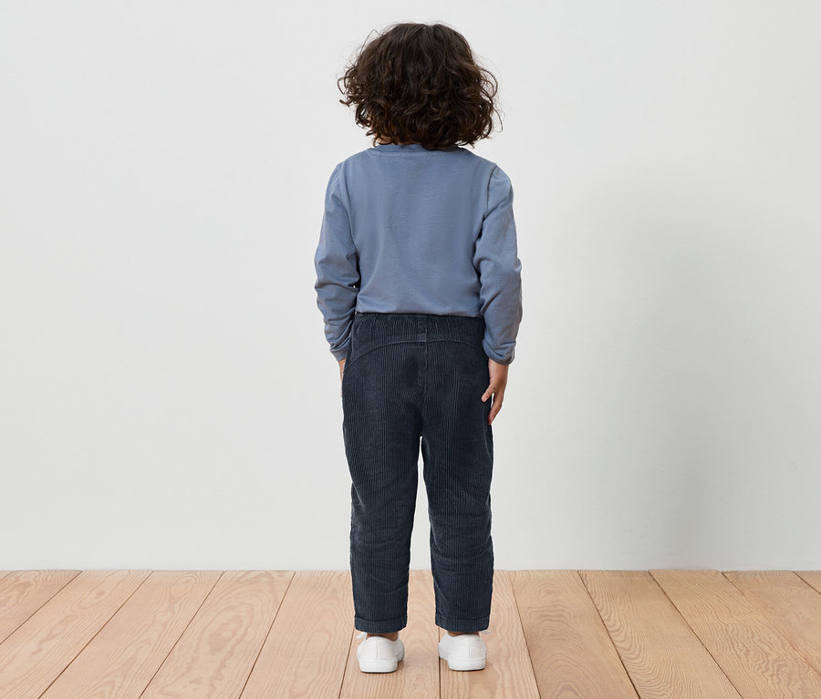 Ein Junge mit braunen lockigen Haaren steht mit dem Rücken zur Kamera und trägt ein blaues Langarmshirt und dunkelblaue Hosen.