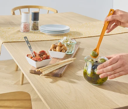 Eine Hand nimmt mit einem orangefarbenen Löffel grüne Oliven aus einem Glas auf einem Holztisch. Auf dem Tisch stehen Schalen mit Tomaten, Pilzen, Käse, Sticks, Salz- und Pfeffermühlen.