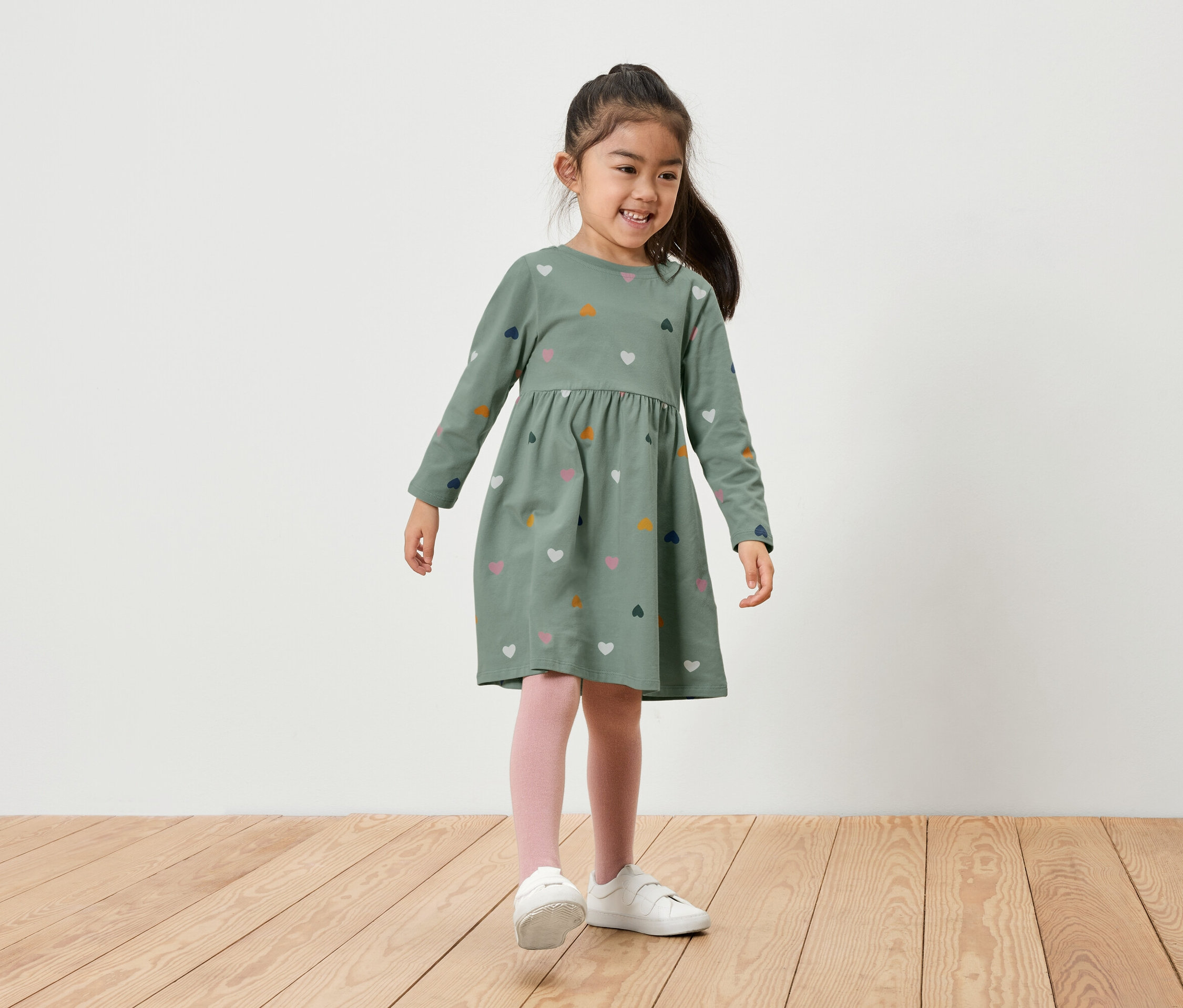 Ein lächelndes Mädchen steht auf einem Holzboden. Sie trägt ein grünes Kleid mit langen Ärmeln und Herzchenmuster, rosa Leggings und weiße Schuhe.
