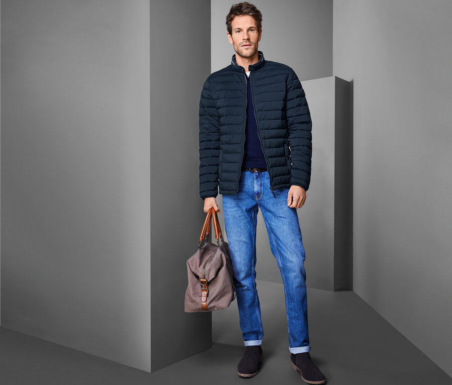 Mann in blauer Steppjacke, Jeans und Schuhen, der eine Tasche hält.