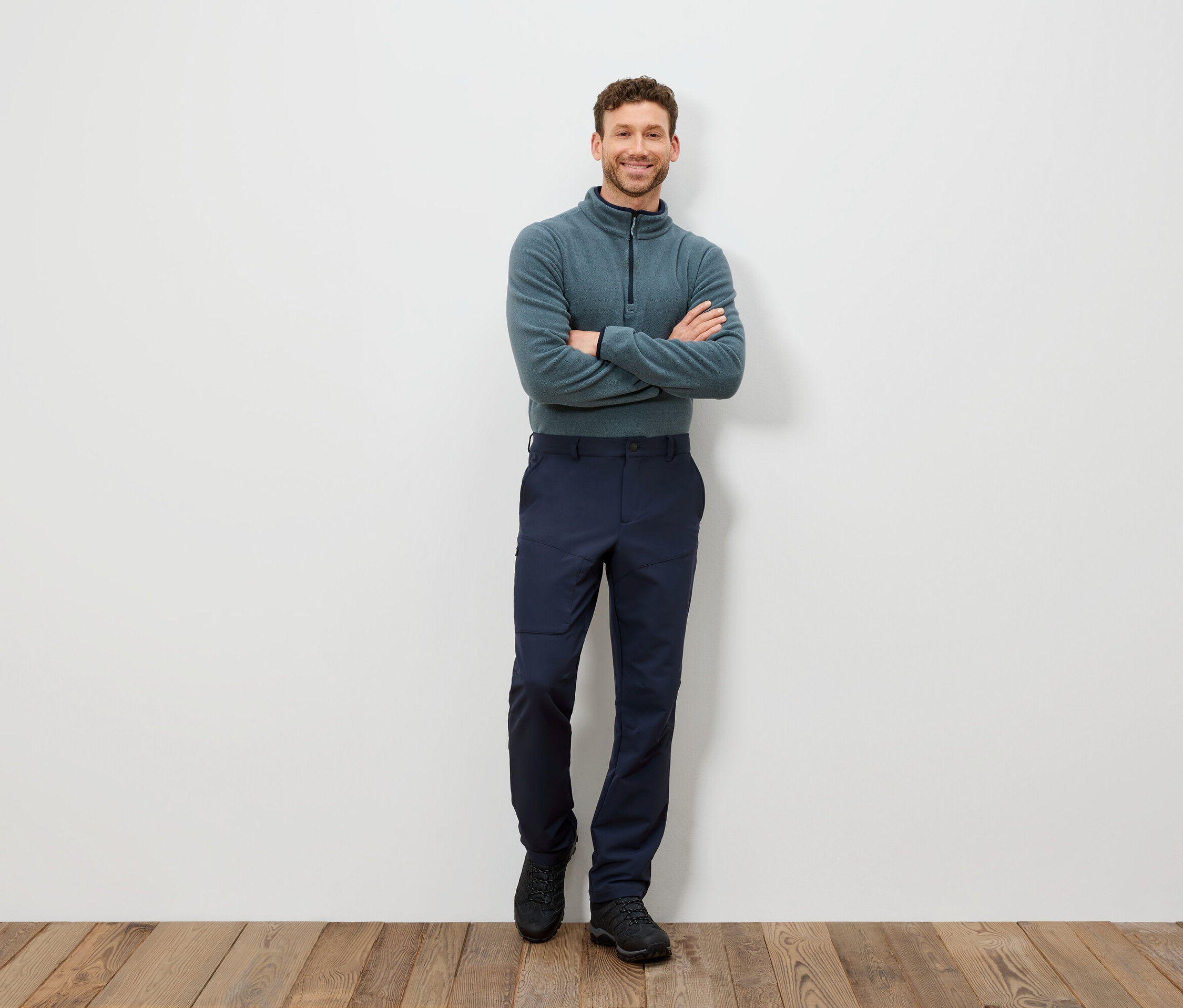 Ein Mann steht mit verschränkten Armen und trägt einen blaugrünen Pullover und dunkelblaue Hosen.