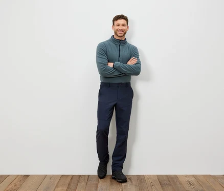Ein Mann steht mit verschränkten Armen und trägt einen blaugrünen Pullover und dunkelblaue Hosen.