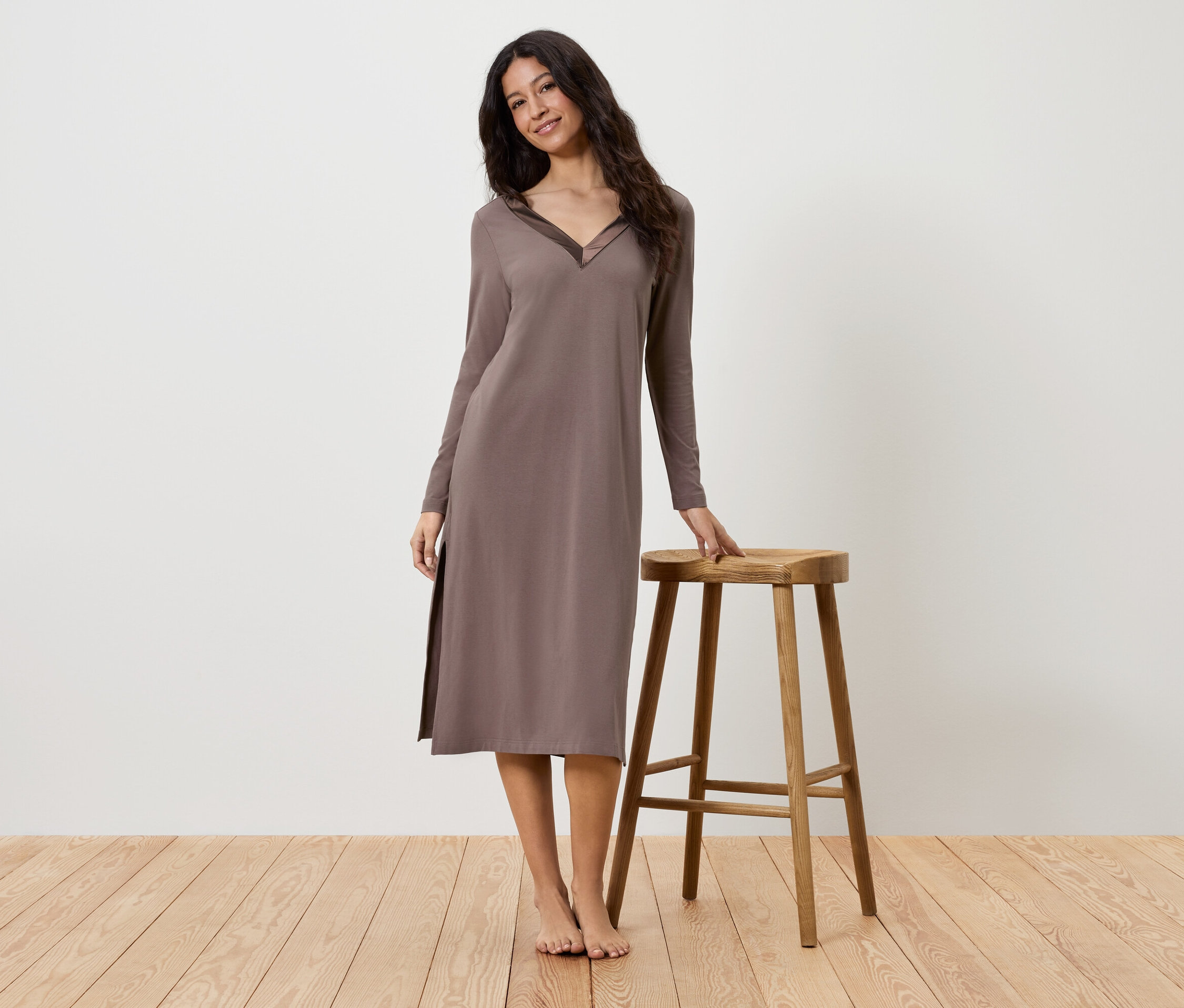 Frau im braunen, langärmligen Kleid, die neben einem Holzhocker steht.