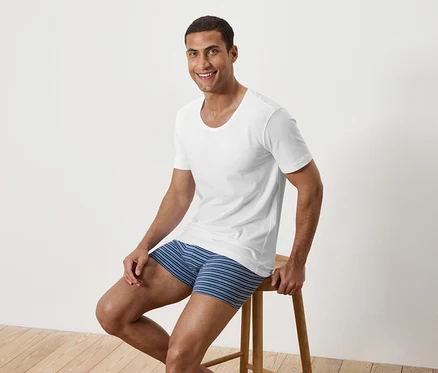 Ein Mann sitzt auf einem Holzhocker, trägt ein weißes T-Shirt und blau gestreifte Boxershorts.