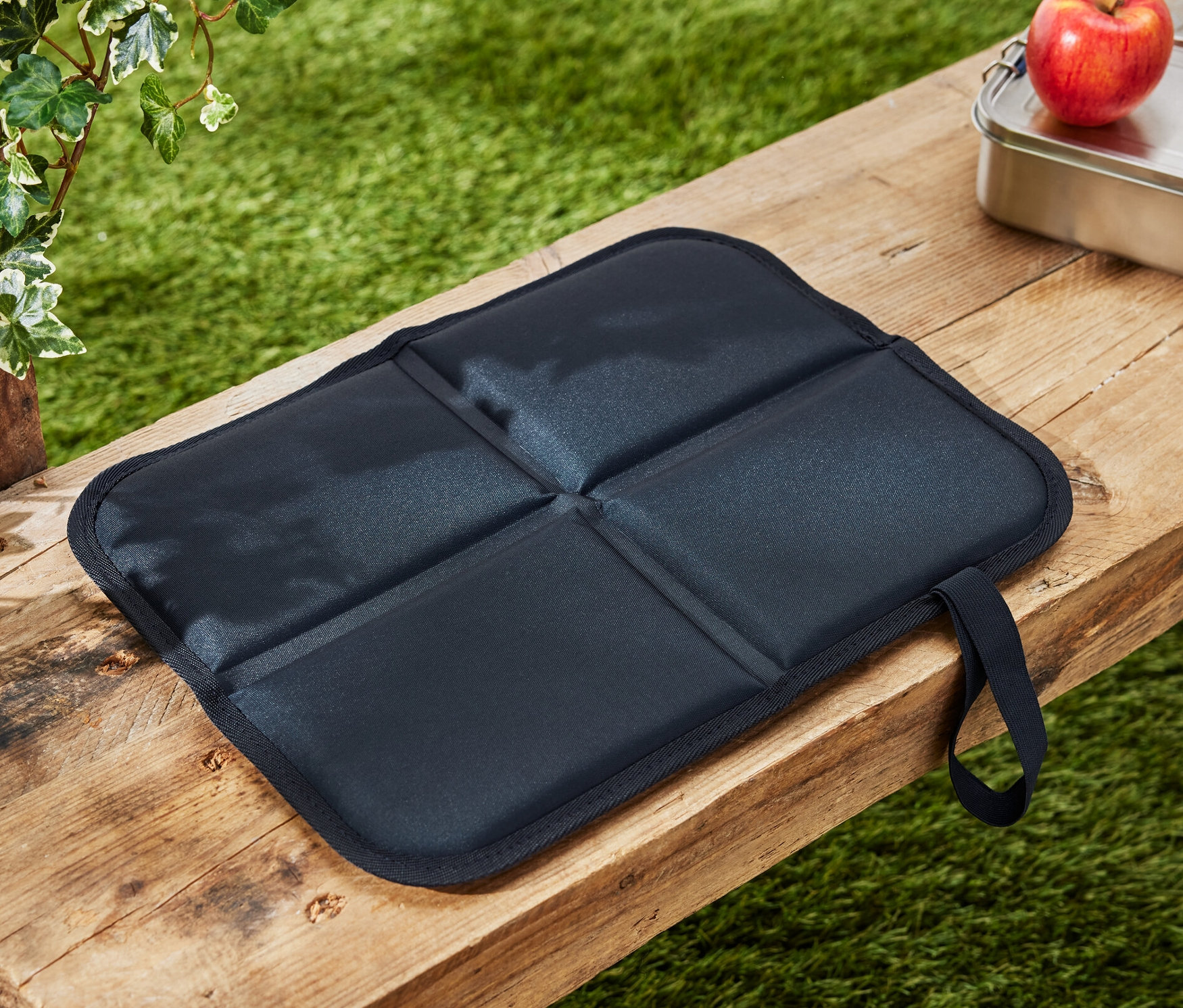 Ein faltbares Thermo-Sitzkissen liegt auf einer Holzbank neben einem Apfel und einer Brotdose.
