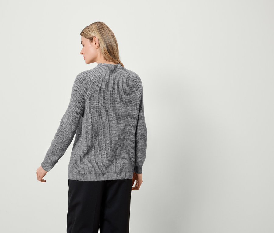 Frau posiert von hinten, trägt einen grauen Pullover und eine schwarze Hose.