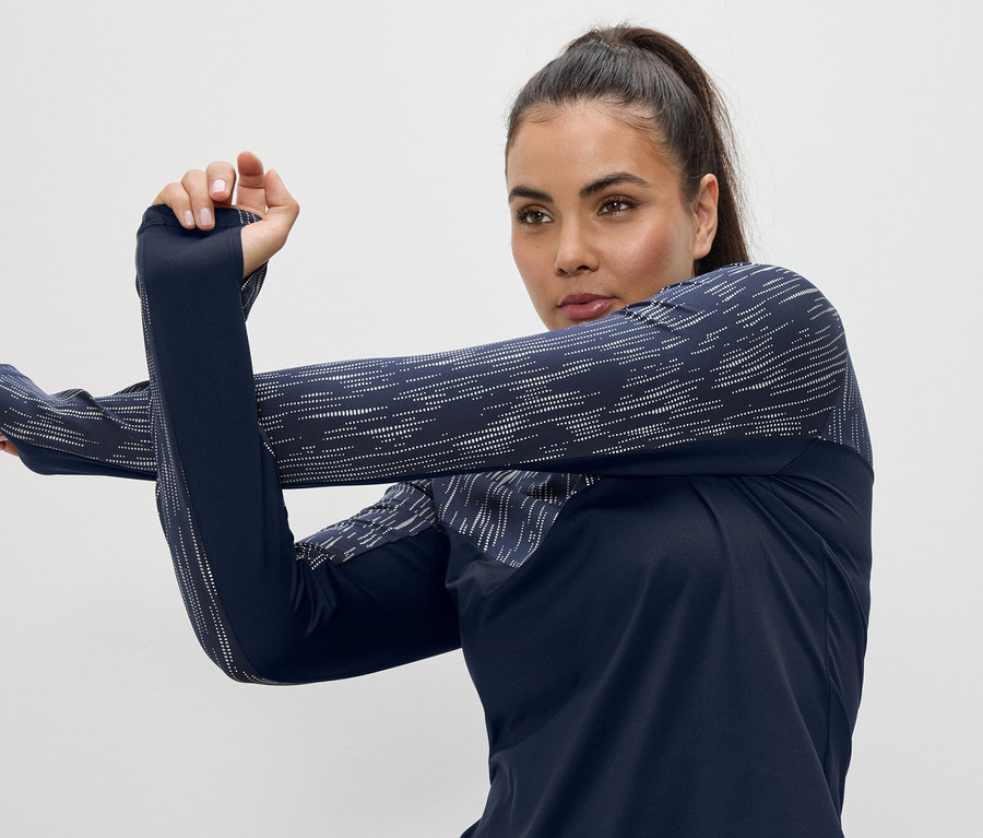 Frau dehnt Arme in einem dunkelblauen, langärmligen Sportoberteil mit Daumenlöchern.