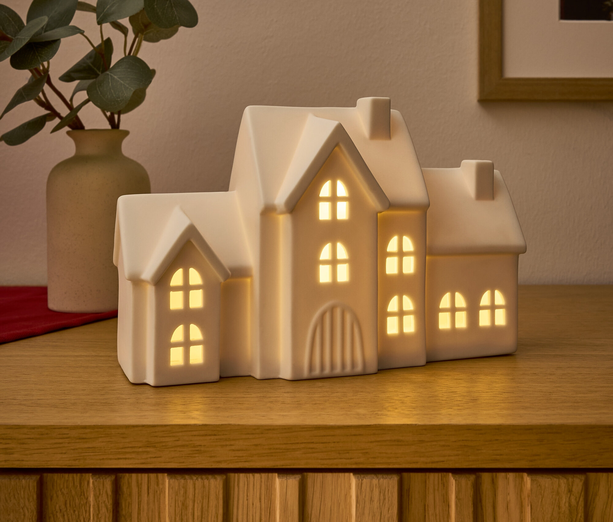 Auf einer Holzkommode stehen ein leuchtendes Keramikhaus und eine Vase mit Eukalyptus.