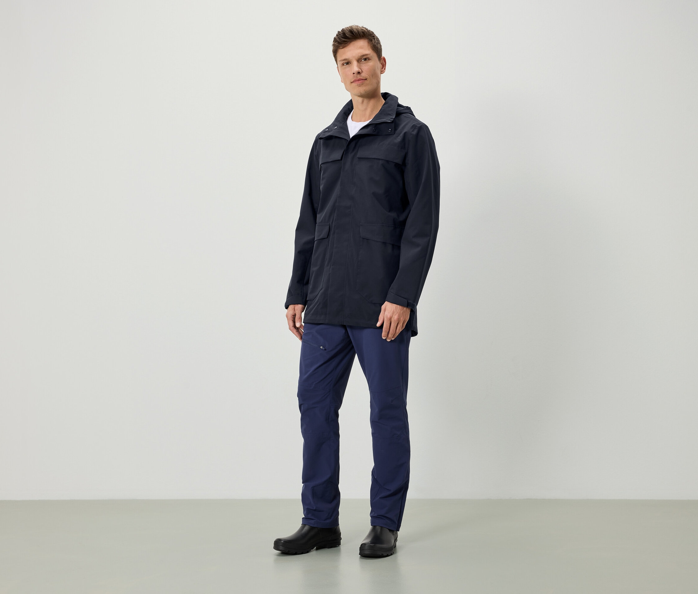 Ein Mann steht und trägt eine dunkelblaue Jacke mit Kapuze, dunkelblaue Hosen und schwarze Schuhe.