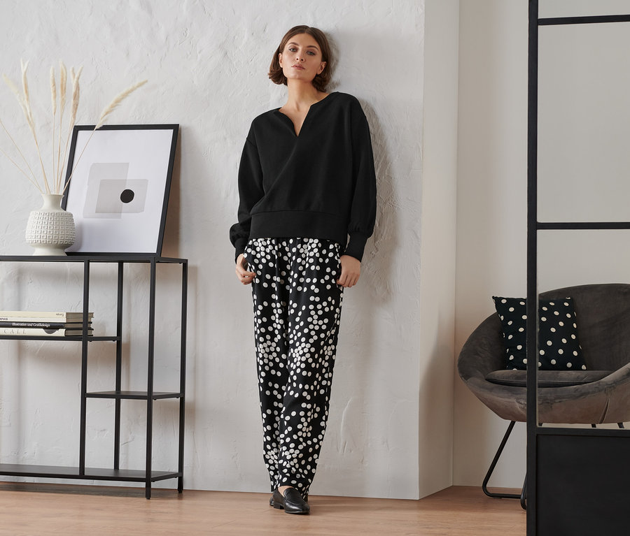 Frau steht und trägt ein schwarzes Sweatshirt und eine schwarz-weiss gepunktete Hose.