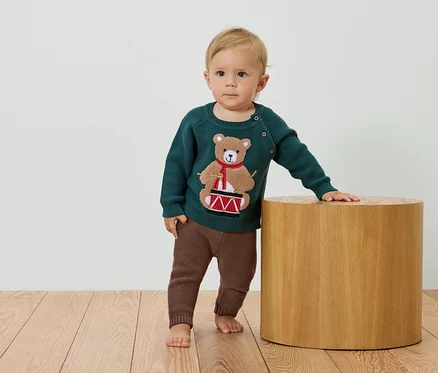 Ein Kind steht neben einem Holzhocker und trägt einen grünen Pullover mit einem Bären, der Trommel spielt, und braune Hosen.