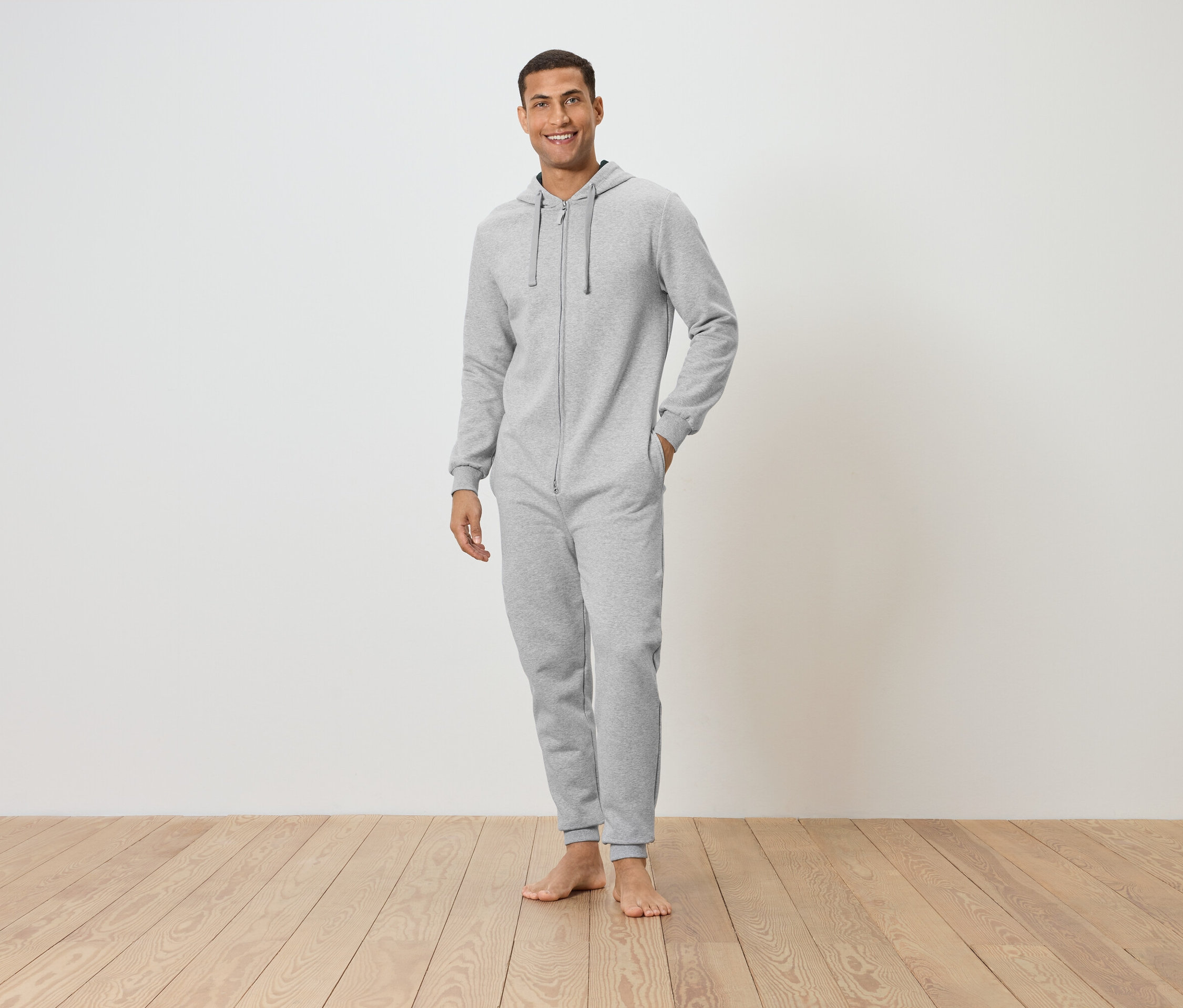 Mann steht barfuß auf Holzboden, trägt einen grauen Overall mit Kapuze und lächelt.
