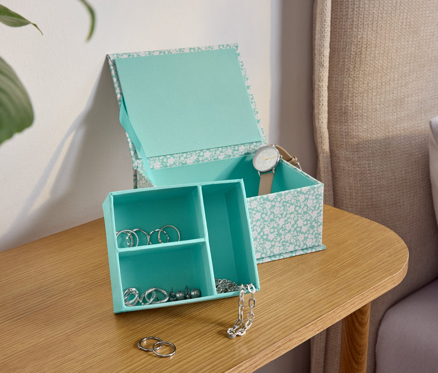 Eine türkisblaue Schmuckschatulle mit Blumenmuster auf einem Holztisch neben einem Bett.