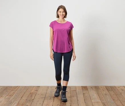 Eine Frau steht auf einem Holzboden vor einer weißen Wand. Sie trägt ein pinkfarbenes T-Shirt, blaue Leggings und graue Stiefel.