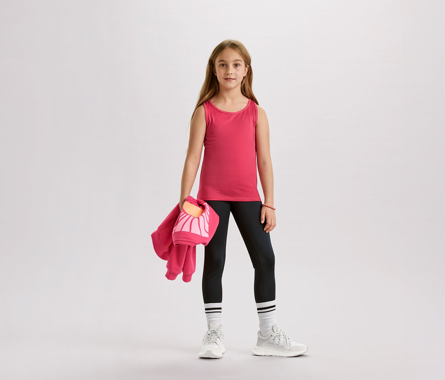 Mädchen mit rosa Tanktop, schwarzen Leggings, weißen Socken und Turnschuhen, hält eine rosa Jacke und eine Tasche.