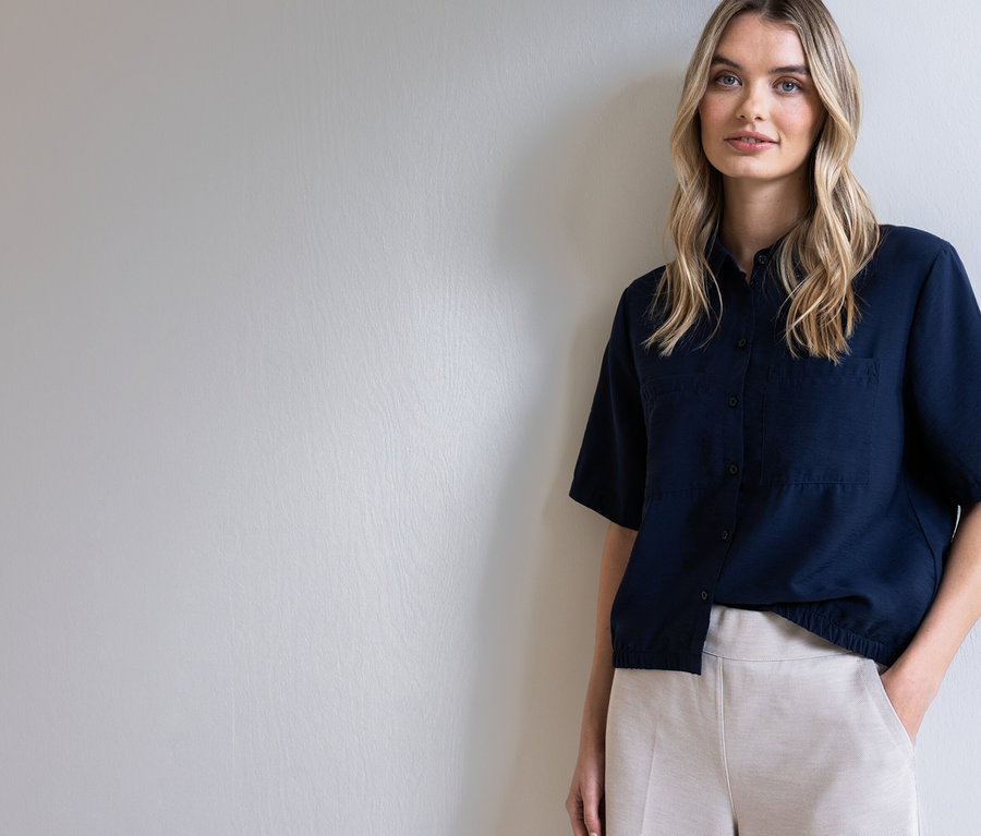 Frau mit blonden Haaren, die ein dunkelblaues Hemd und beigefarbene Hosen trägt, posiert vor einer hellgrauen Wand.