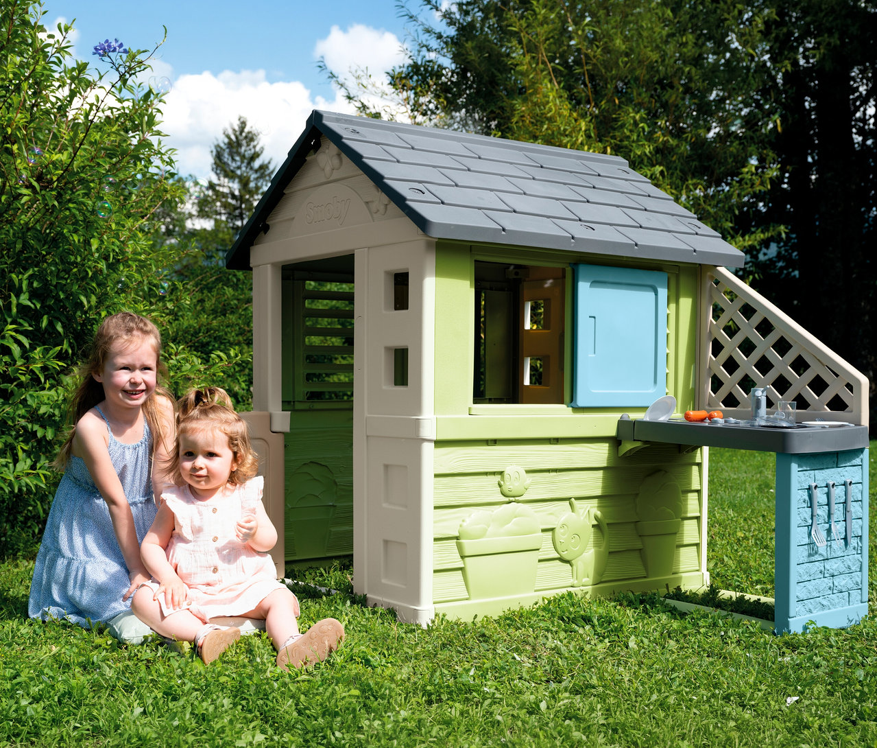 Zwei Mädchen sitzen vor dem Smoby Life Spielhaus »Pretty« mit Küche im Garten.