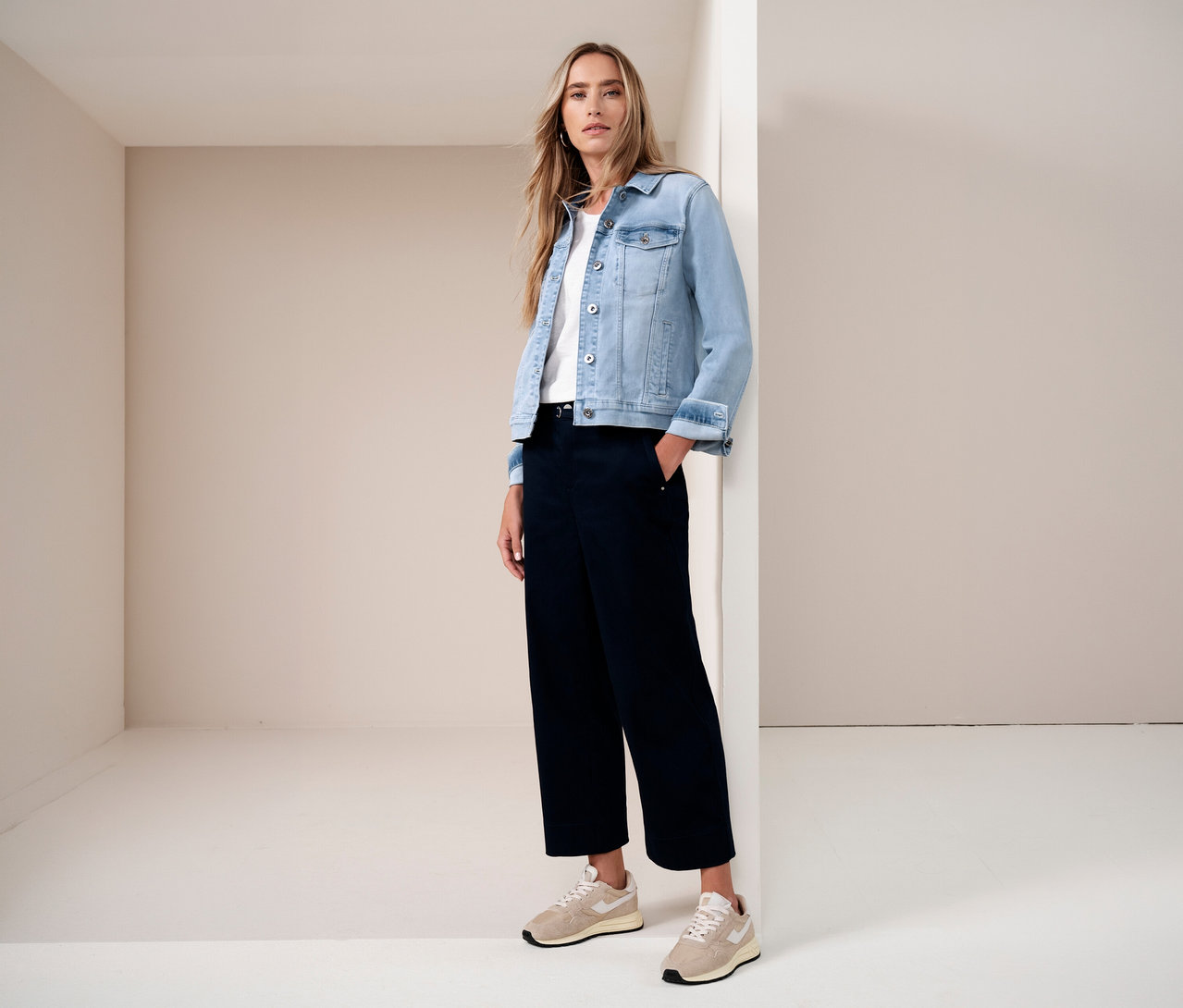 Frau in Jeansjacke, weißem T-Shirt und dunkler Hose, mit einer Hand in der Hosentasche, posiert vor einer Ecke in einem hellen Raum.