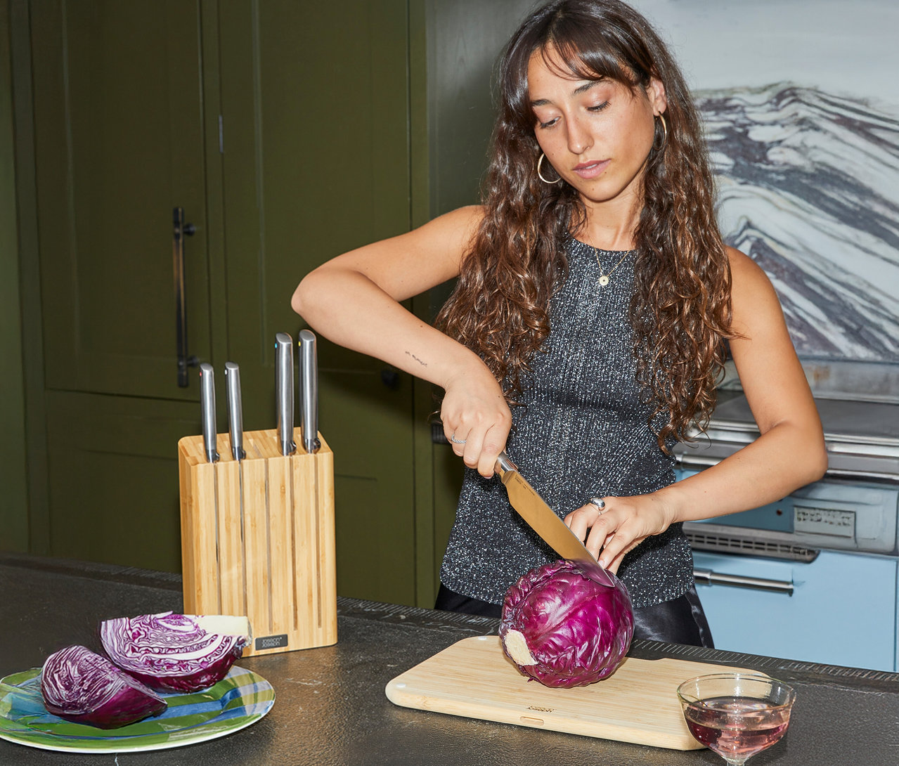 Frau schneidet Rotkohl auf einem Schneidebrett. Ein Messerblock und ein Glas mit einem rosa Getränk stehen auf der Küchentheke.