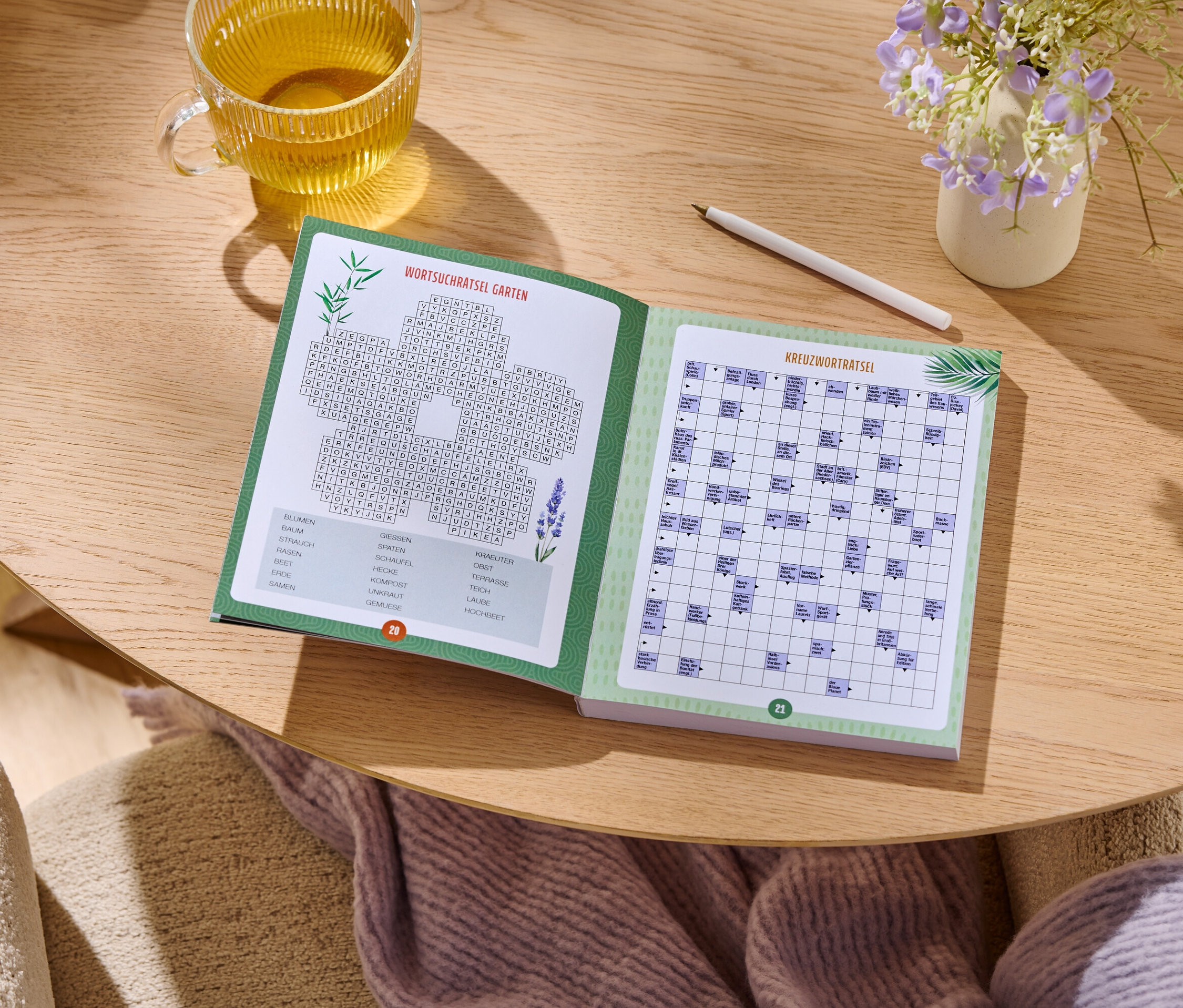 Auf einem Holztisch liegt ein aufgeschlagenes Rätselbuch neben einem Glas Tee, einem Stift und einer kleinen Vase mit Blumen.