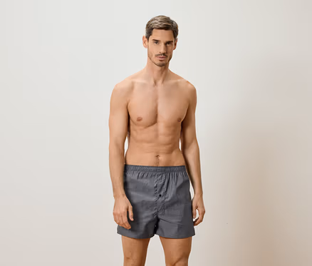 Mann mit freiem Oberkörper, der graue Boxershorts trägt und vor einem hellen Hintergrund steht.