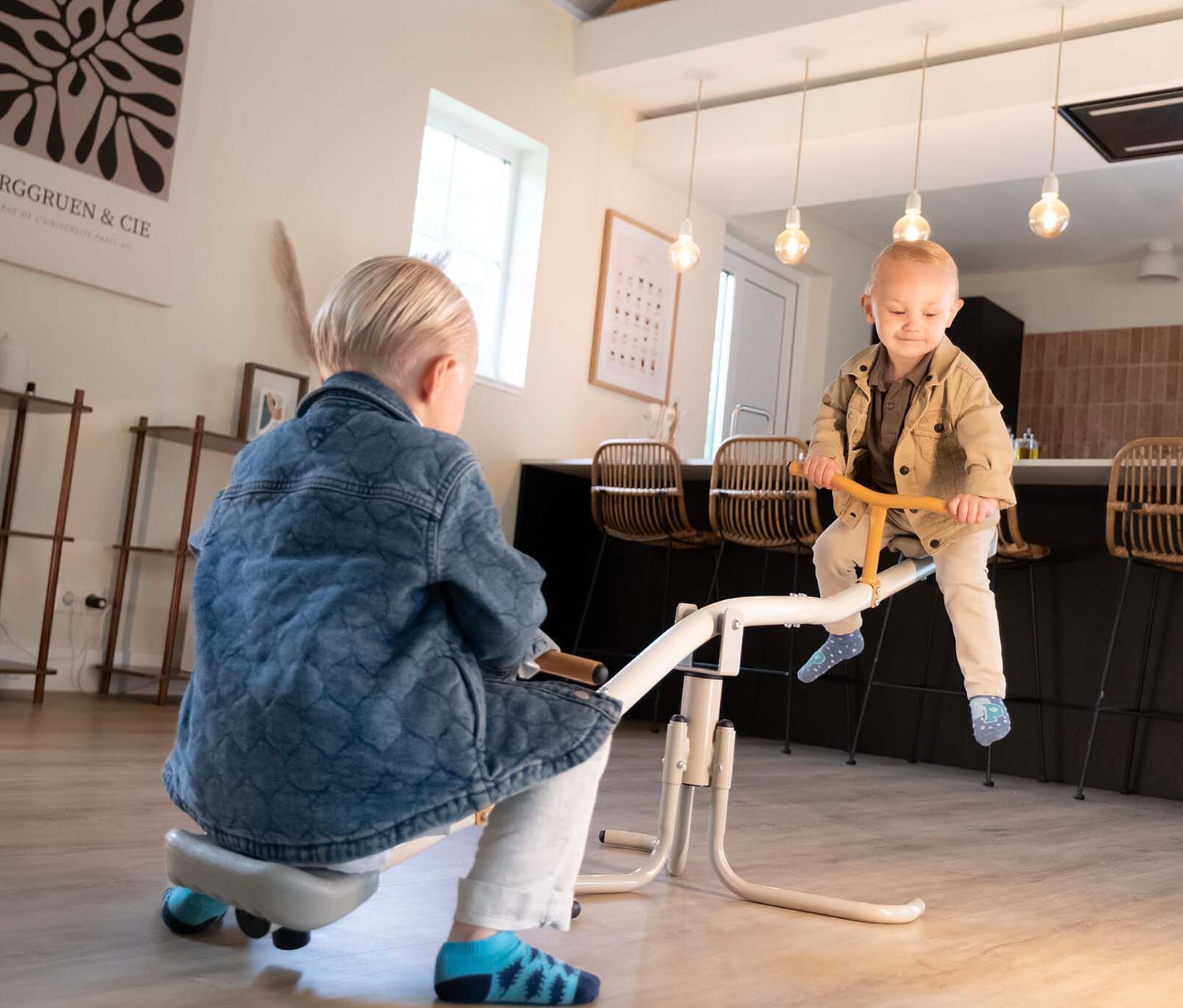 Zwei Kinder spielen auf einer weißen Wippe in einem hellen Raum mit Holzboden.