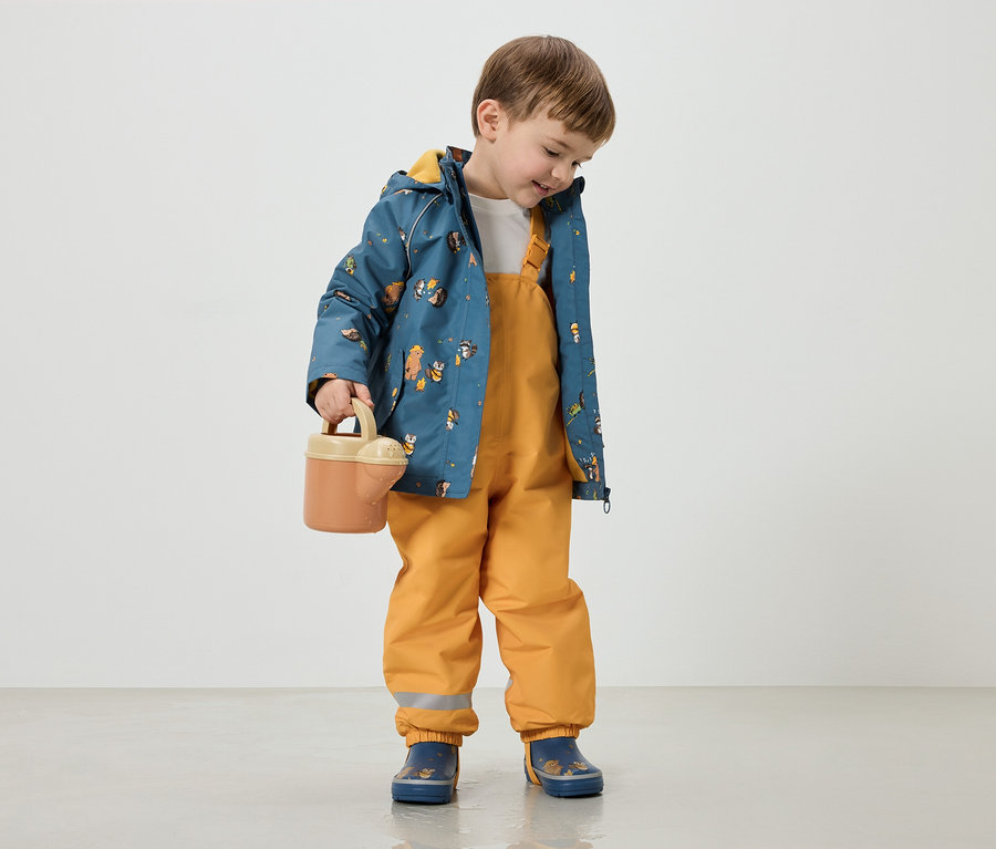 Ein Junge mit braunen Haaren trägt eine blaue Jacke mit Tiermotiven, gelbe Latzhose und blaue Stiefel und hält eine Gießkanne.
