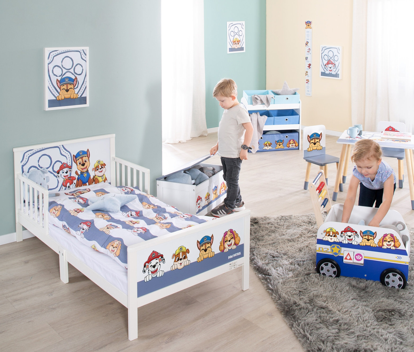 Ein Kinderzimmer mit einem Jungen, der an einer Spielzeugkiste steht, und einem Mädchen, das in einer Paw Patrol Spielzeugkiste spielt.