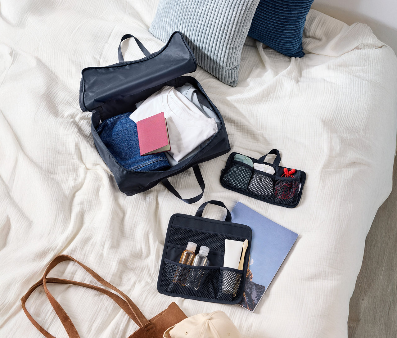 Draufsicht auf einen Koffer mit Kleidung, zwei Kissen, eine Kosmetiktasche, eine Tragetasche und eine Kappe auf einem Bett.